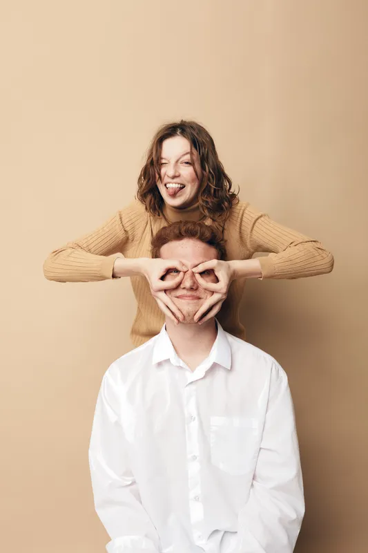 Happy young woman in beige sweater playfully covers eyes of smiling man in white shirt. Studio portrait on beige background. Concept of friendship, fun, joy, and playful relationship moments.