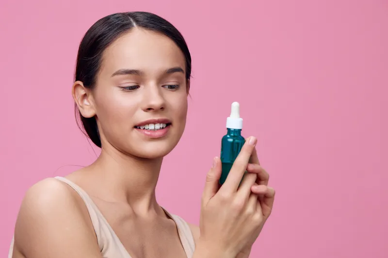 Young woman holding a skincare serum bottle, expressing joy against a pink background, showcasing beauty and self care concepts for healthy skin