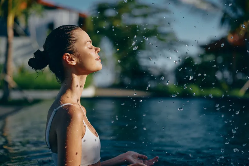 A woman in a white swimsuit stands by a pool with eyes closed, basking in warm sunlight. Water droplets spray around, capturing a moment of outdoor relaxation.