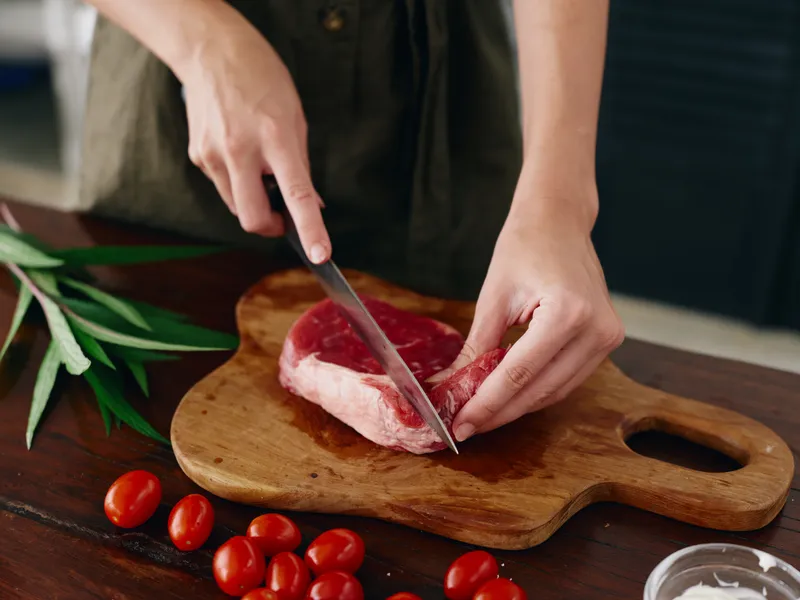 Женщина с ножом в руке нарезает свежий стейк для запекания на кухне