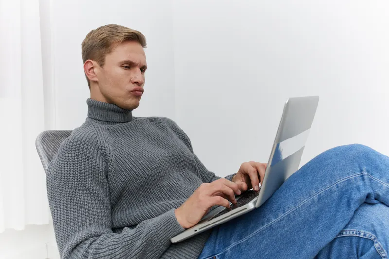 Man working on laptop computer while sitting on a couch in a modern minimalistic room, casual clothing, focused expression, technology concept, indoor lifestyle, relaxing at home, using digital device
