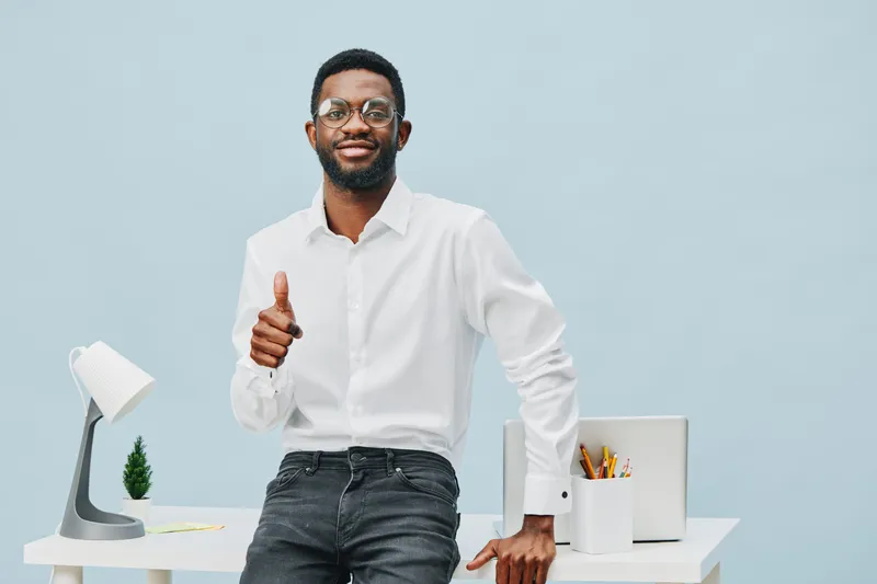 Confident smiling young African American man in white shirt and dark jeans standing by desk with office supplies and modern lamp, cheerful and professional indoor portrait for business or workplace co