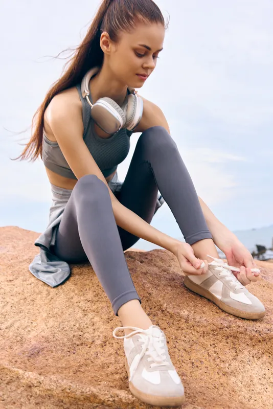 Athletic young woman tying shoelaces on a rock, wearing stylish workout clothing, headphones, showing a focused expression, with a natural outdoor background