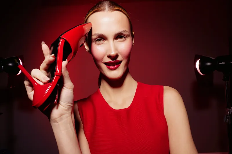 Smiling woman in red dress holds red high heel shoe near face against red background. Fashion concept with confident model showcasing stylish footwear and elegant look in studio lighting.