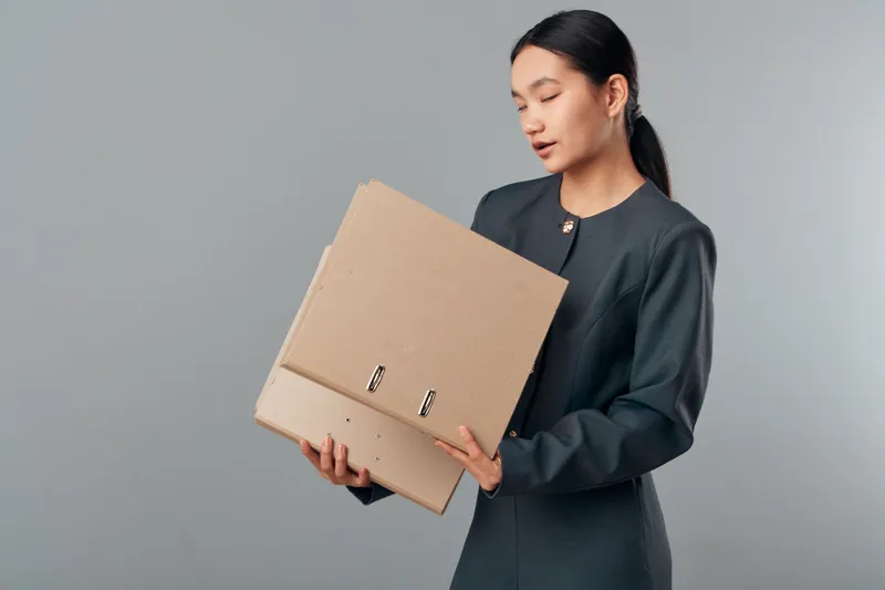 Woman holding cardboard folders for office documents and paperwork standing against gray background in formal business attire. Female employee organizing files for work task.