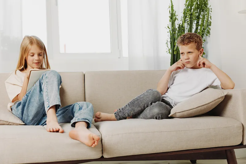 Two children sit on a beige sofa in a bright living room, one girl focused on a tablet while a boy covers his ears showing frustration. Concept of technology, emotions, and family life.