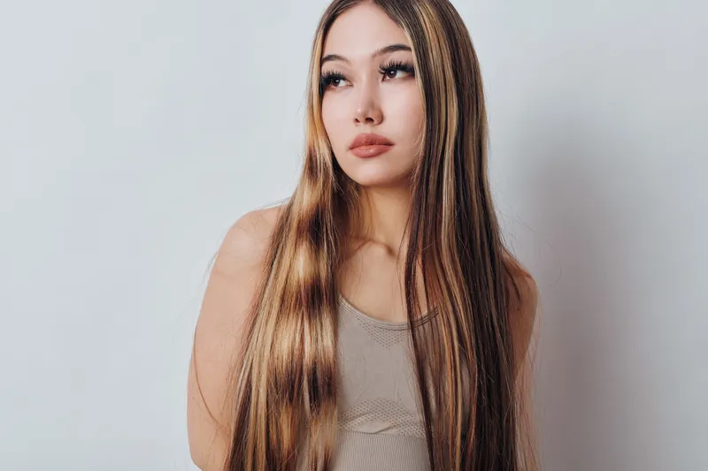 Young woman with long straight hair and natural makeup wearing casual sleeveless top standing against plain white background looking sideways.