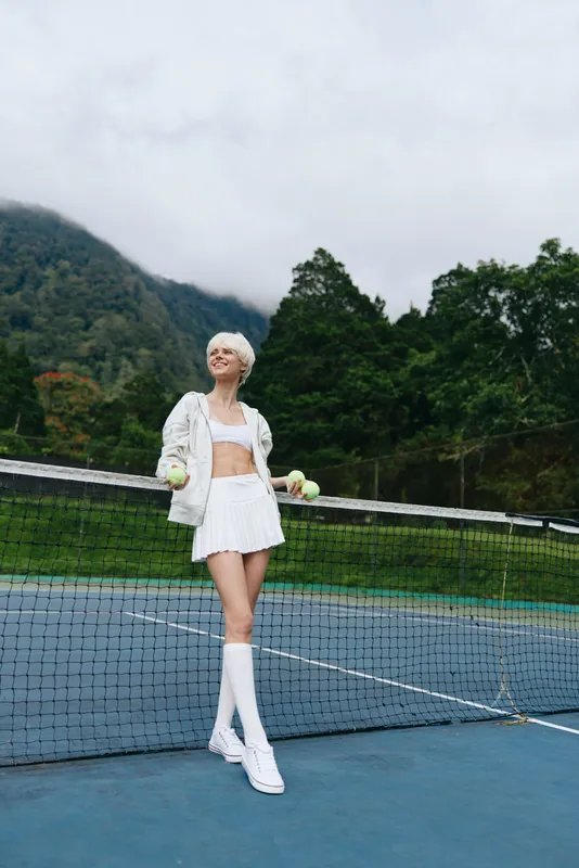 Young athletic woman in sporty white outfit, holding tennis balls, smiling on outdoor tennis court with green background and cloudy sky, embodying active lifestyle and confidence