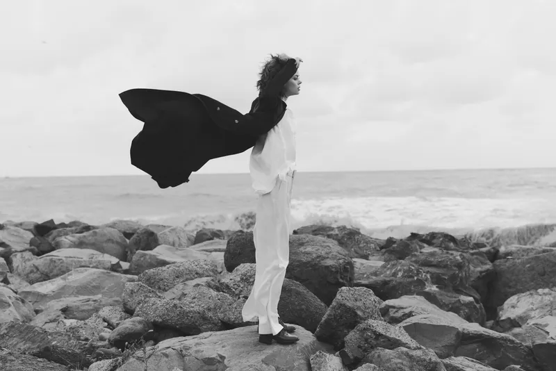 Woman stands on rocky coastline with a flowing cape, white attire contrasting the gray sea, wind shaping motion and calm stance for a dramatic, timeless coastal scene