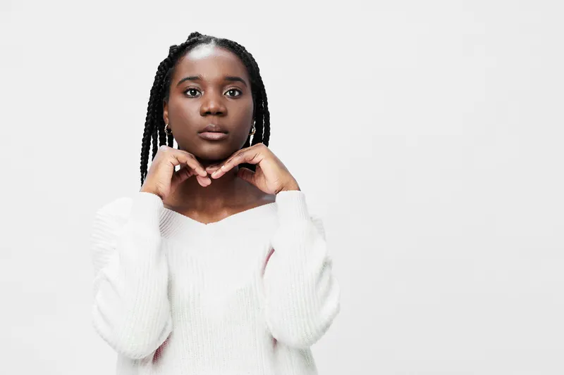 A young Black woman wearing a cozy white sweater poses thoughtfully against a plain background, expressing a serene yet contemplative emotion