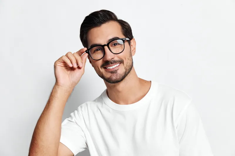 Smiling man wearing glasses and a white t-shirt, looking at the camera while adjusting his glasses with a cheerful expression against a plain white background.