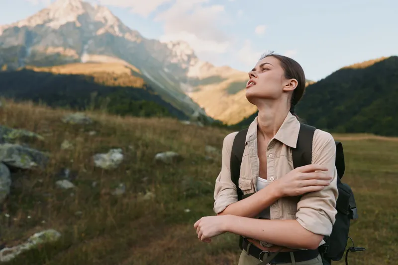 A woman outdoors with a backpack stands among mountains, eyes closed, face lifted to the sun, embodying freedom, resilience, and personal discovery.