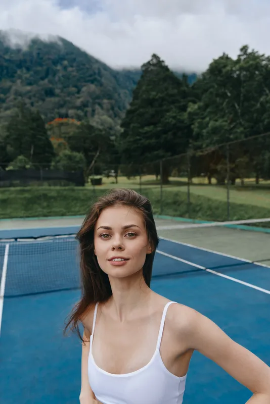 Young woman in sporty attire smiling on a tennis court surrounded by lush greenery and mountains, conveying a vibrant and active lifestyle