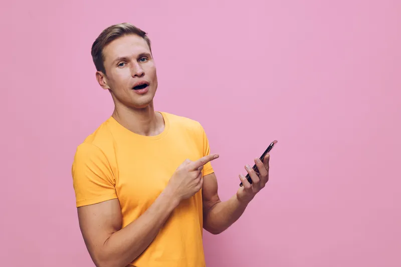 Young man in casual yellow t-shirt pointing at smartphone with surprised expression against pink background. Studio portrait of a person showing curiosity and surprise. Modern lifestyle concept.