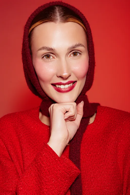 Smiling woman with red lips and rosy cheeks wears red sweater and matching headscarf, posing with hand near chin on red background. Concept of beauty, fashion, and winter style.
