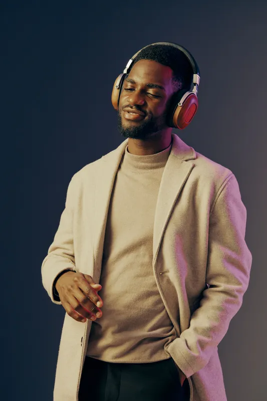 Smiling young Black man in beige coat and headphones enjoying music with vibrant background, conveying emotions of relaxation and joy