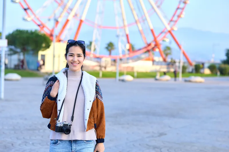Woman with camera exploring park amusement, rollercoaster backdrop, outdoor leisure, smile, photography vibe, traveler styling casual jacket, denim and sweater, candid lens captures vibrant scene