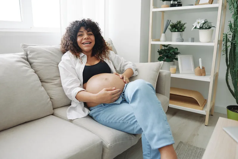 Беременная женщина улыбается и счастливо лежит на диване, чувствуя свободу, и гладит свой живот с ребёнком на последнем месяце беременности