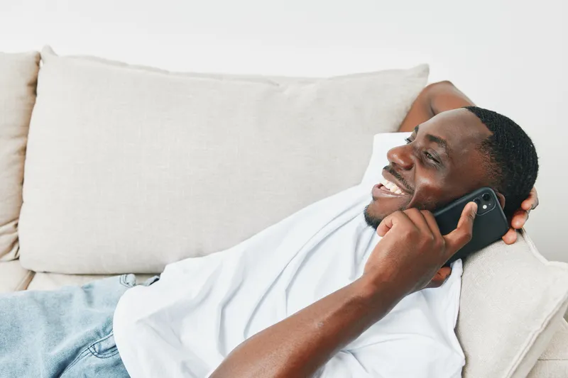 Young Black man enjoying a phone call while relaxing on a sofa, wearing a casual white t shirt, with a joyful expression and light background