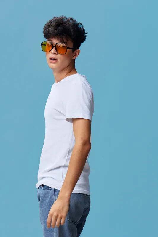 young man wearing yellow sunglasses and white t-shirt standing against blue background with casual pose and curly hair style portrait