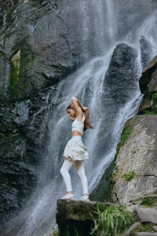 Active woman stretching near waterfall in white fitness clothes, surrounded by rocky nature and lush greenery, showcasing outdoor exercise and wellness.