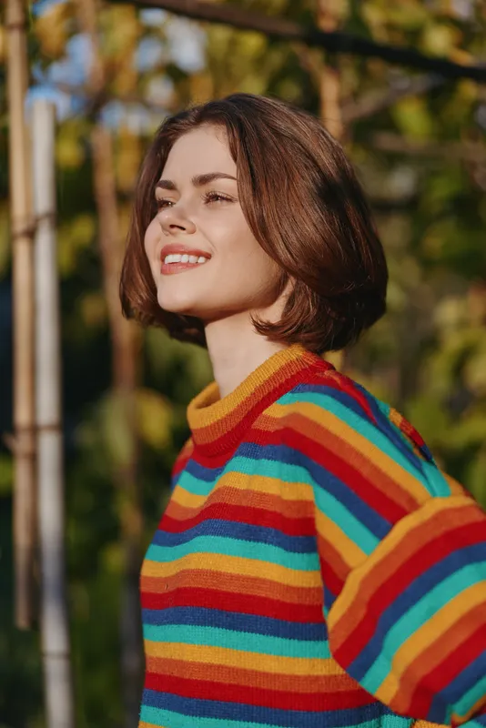 Young woman in her 20s wearing a colorful striped sweater, smiling and enjoying warm sunlight outdoors. Casual bob haircut, relaxed lifestyle portrait in nature.