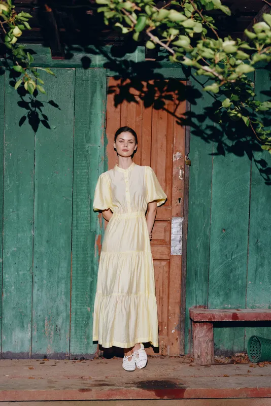 A girl in a pale yellow dress stands in front of a green wooden doorway outdoors, creating a vivid portrait that blends fashion charm with casual rustic atmosphere and natural light