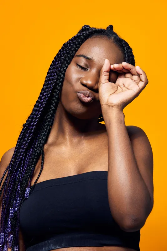Confident young Black woman with braided hair playfully making a gesture against a vibrant yellow background, showcasing bold style and positive vibes