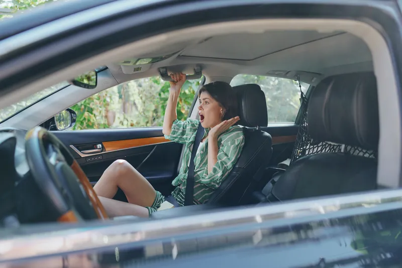 Young woman in a car shows fear and surprise, gripping the headrest while seated in the driver's seat.