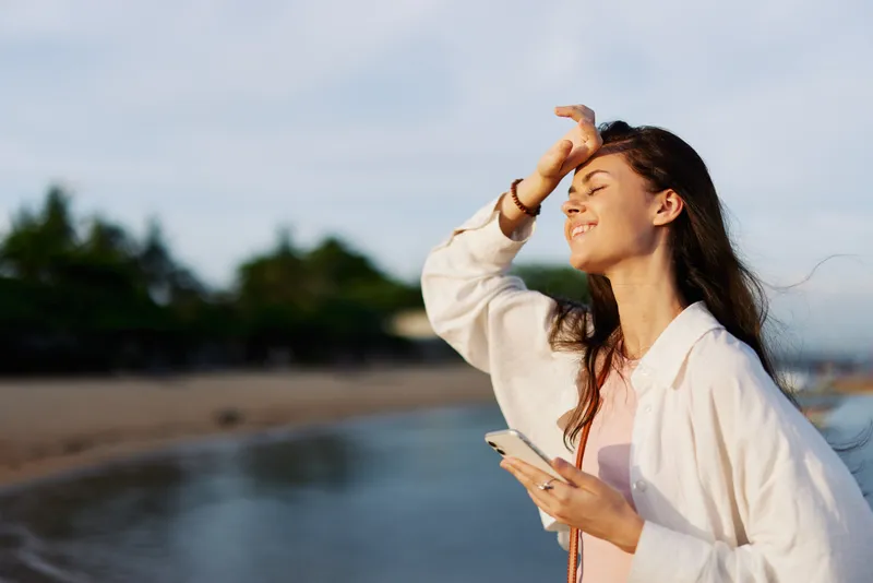 Женщина-фрилансер улыбается с телефоном в руке на отдыхе, гуляя по пляжу у океана на Бали