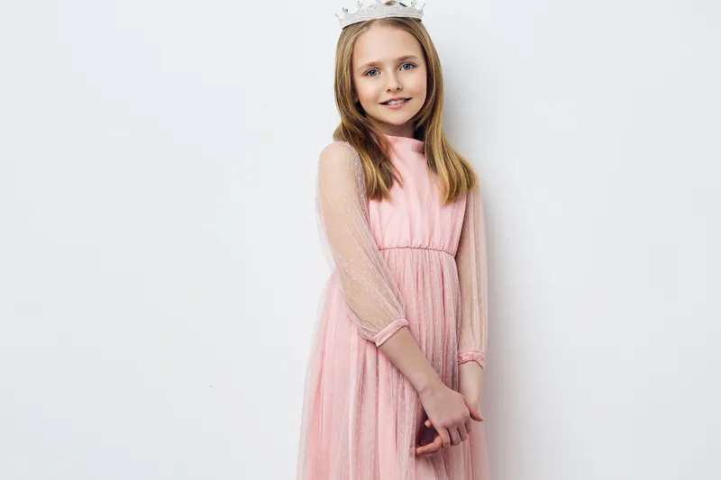 Charming young girl in a pink dress wearing a crown, smiling happily in a bright and airy studio setting, perfect for children s events and celebrations