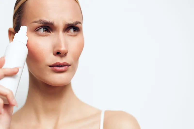 Closeup of a woman applying a white skincare product to her cheek, highlighting a skincare routine, hydration, and glow on a neutral background. Focus on selfcare, beauty, and facial care.