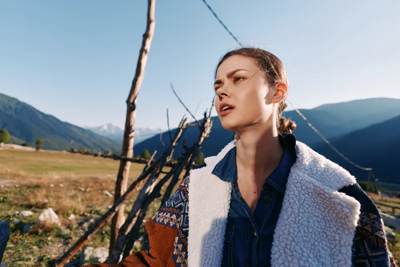 Woman portrait in mountains countryside outdoors wearing a warm jacket, sunlight on her face, young traveler near a wooden fence with scenic landscape and clear blue sky above.