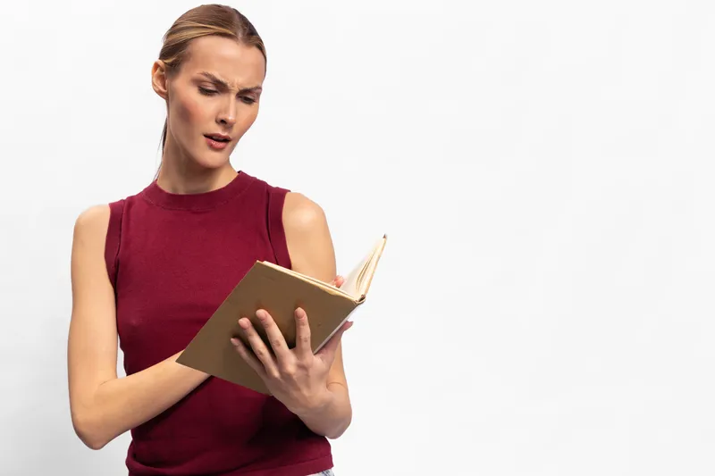 Confused young woman in red sleeveless top reading a notebook with focused expression. Isolated on white background. Concept of concentration, study, and decision making.