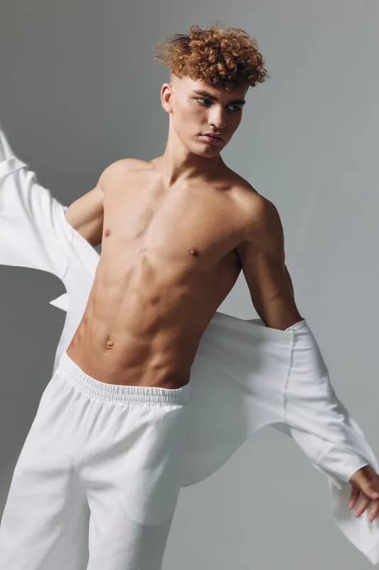 Young man with curly hair and toned body wearing white pants and removing white shirt in studio. Muscular male model posing with focused expression on gray background.