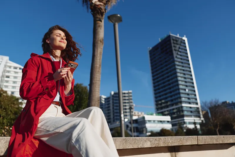 Young woman in a red coat sits on a ledge in an urban park, smiling while holding a smartphone, with modern highrise buildings and a bright blue sky behind.