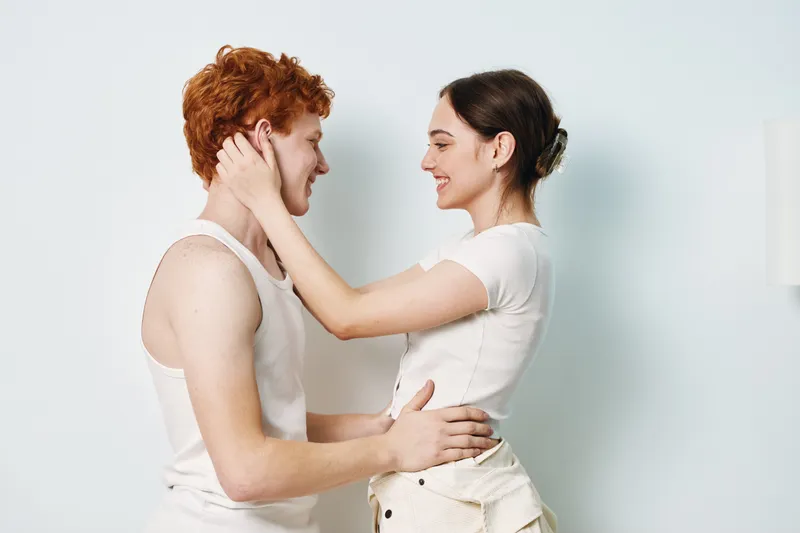 Couple, young man and woman in white clothing, smiling and embracing with tenderness in light room. Intimacy, love, connection and happiness between two people with gentle touch.