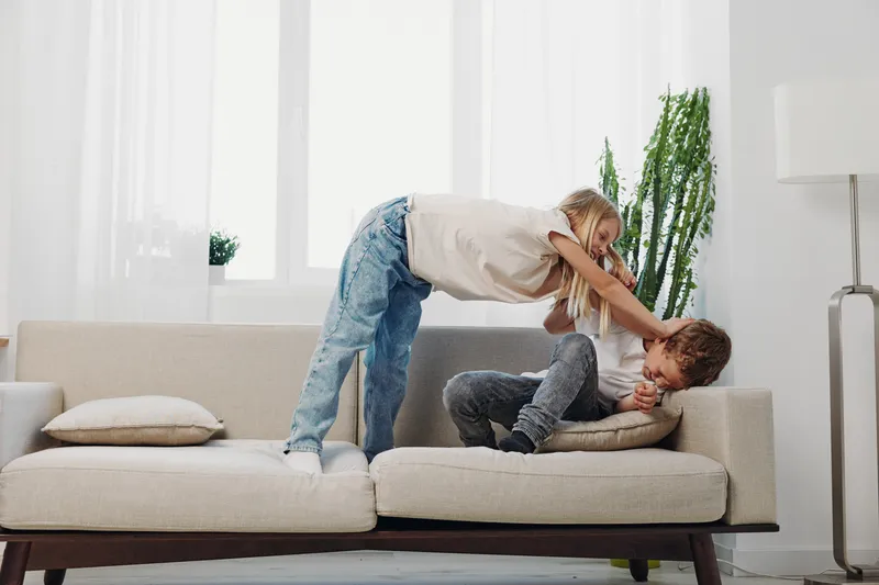 Playful children having fun on sofa in bright living room, girl tickling boy who laughs joyfully. Concept of childhood, family time, happiness, and indoor activity.
