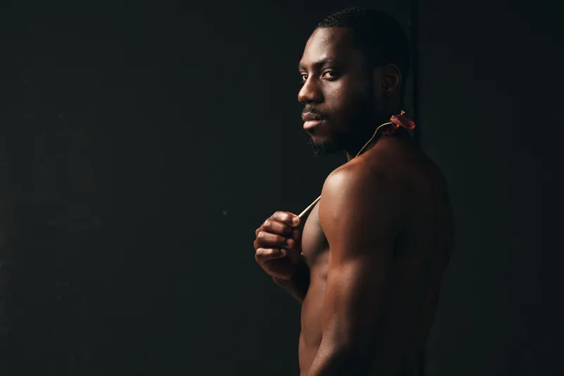African man with dark skin in creative conceptual style posing shirtless with flower on shoulder against dark background for artistic portrait and visual expression.