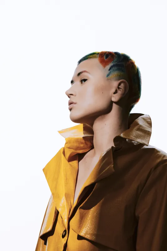 Bold woman with colorful shaved hair and yellow jacket, posing confidently against a bright white background in a modern avant-garde style portrait.