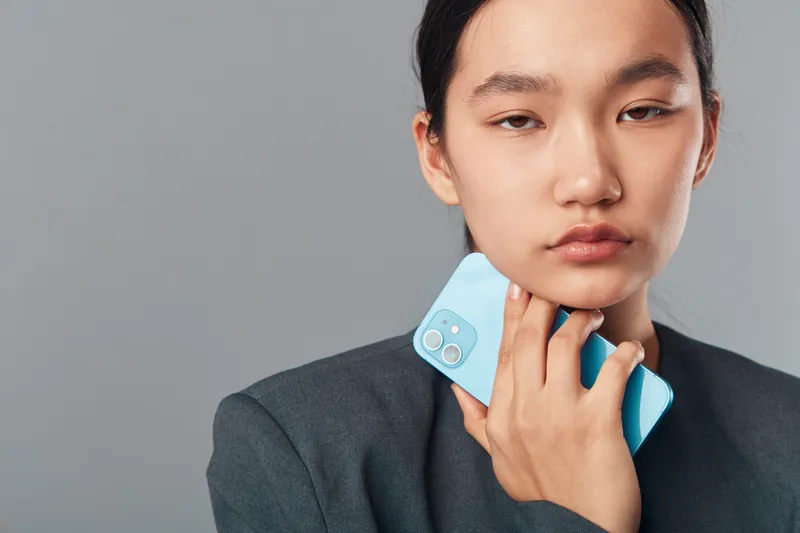 Young woman holding light blue smartphone close to face in thoughtful expression with gray background. Casual portrait with soft natural lighting and minimalistic style.