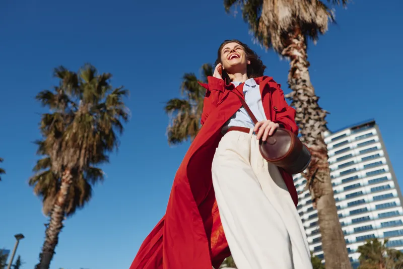 A smiling woman in a red coat stands outdoors under a clear blue sky, palm trees and a modern building behind. Concept for bold fashion and sunny confidence.