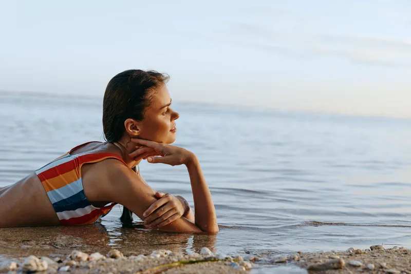 Woman relaxing in shallow water at the beach, striped swimsuit and bare shoulders, looking to the horizon by the ocean, summer sunset light, calm mood, soft waves, warm air, peaceful smile