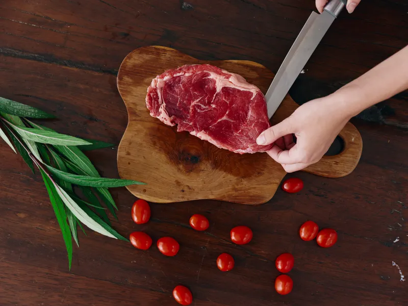 Женщина с ножом в руке нарезает свежий стейк из мяса для жарки на кухне