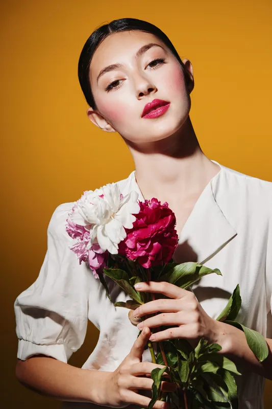 Young woman with colorful fresh flowers holds bouquet posing on yellow background. Vibrant makeup and stylish white dress create fashion portrait. Concept of beauty creativity.