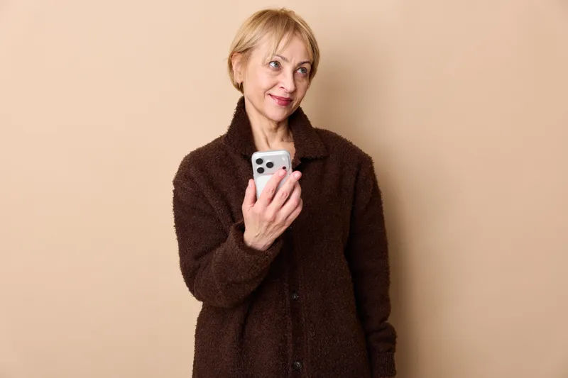 Woman with smartphone in studio portrait, smile on her face, beige background, short blond hair, warm expression. Everyday technology and communication idea, concept.