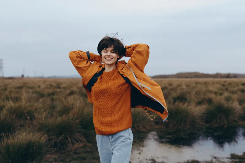 Woman outdoors, smiling in nature field, casual lifestyle portrait, hands behind head pose, standing by river, cloudy overcast light, relaxed cheerful mood