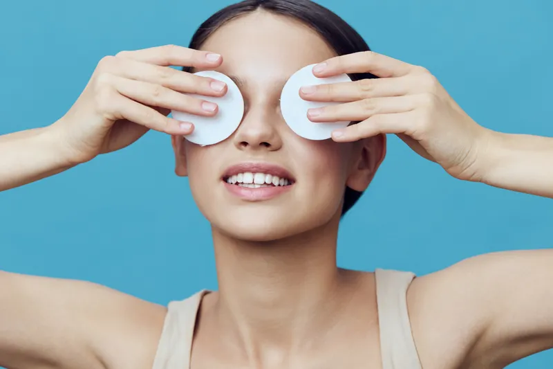 Young woman enjoying skincare routine with cotton pads on her eyes, smiling against a light blue background, conveying relaxation and self care
