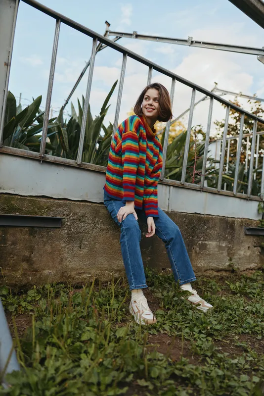 woman striped sweater jeans sitting smiling urban garden: young woman in her 20s wearing a colorful striped sweater and jeans, sitting on a low wall in an urban garden, smiling in white sneakers and