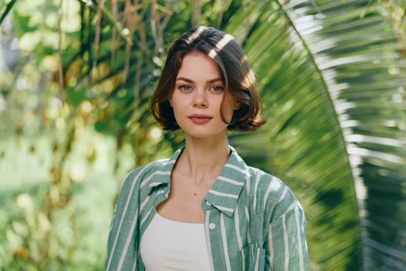 Portrait of a young woman in a tropical garden, wearing a green striped shirt over a white top, bathed in soft sunlight.